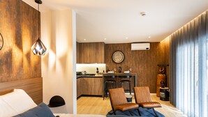 Une chambre d’hôtel moderne comprenant un lit, un coin salon avec deux fauteuils, une petite kitchenette et une grande fenêtre avec des rideaux.