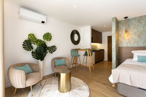 Una habitación de hotel moderna con una cama, dos sillones, una pequeña mesa redonda, una planta en maceta, una zona de cocina y un aire acondicionado de pared.