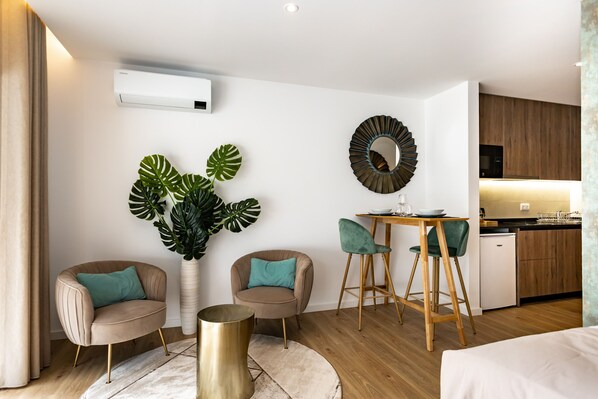 Una sala de estar moderna con un sofá, sillas, una mesa de centro, una planta grande y una cocina con armarios de madera y un refrigerador.