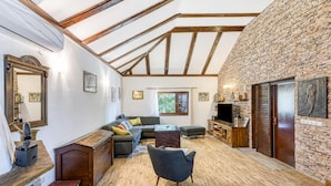 Een ruime woonkamer met een hoge plafond, zichtbare balken, een stenen muur, een open haard en een televisie.