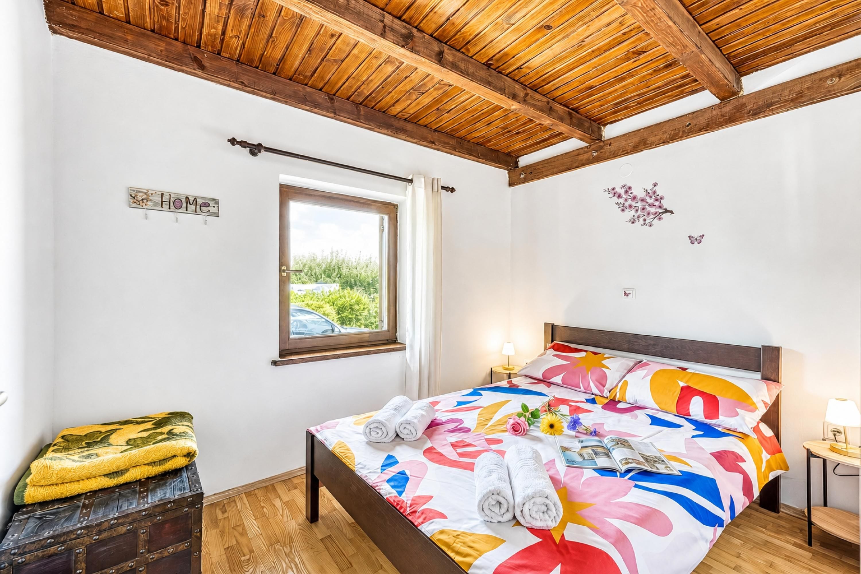 Une chambre avec un plafond en bois, un lit recouvert d’une courtepointe à motifs floraux colorés, une fenêtre donnant sur la verdure et une petite table de chevet avec une lampe.