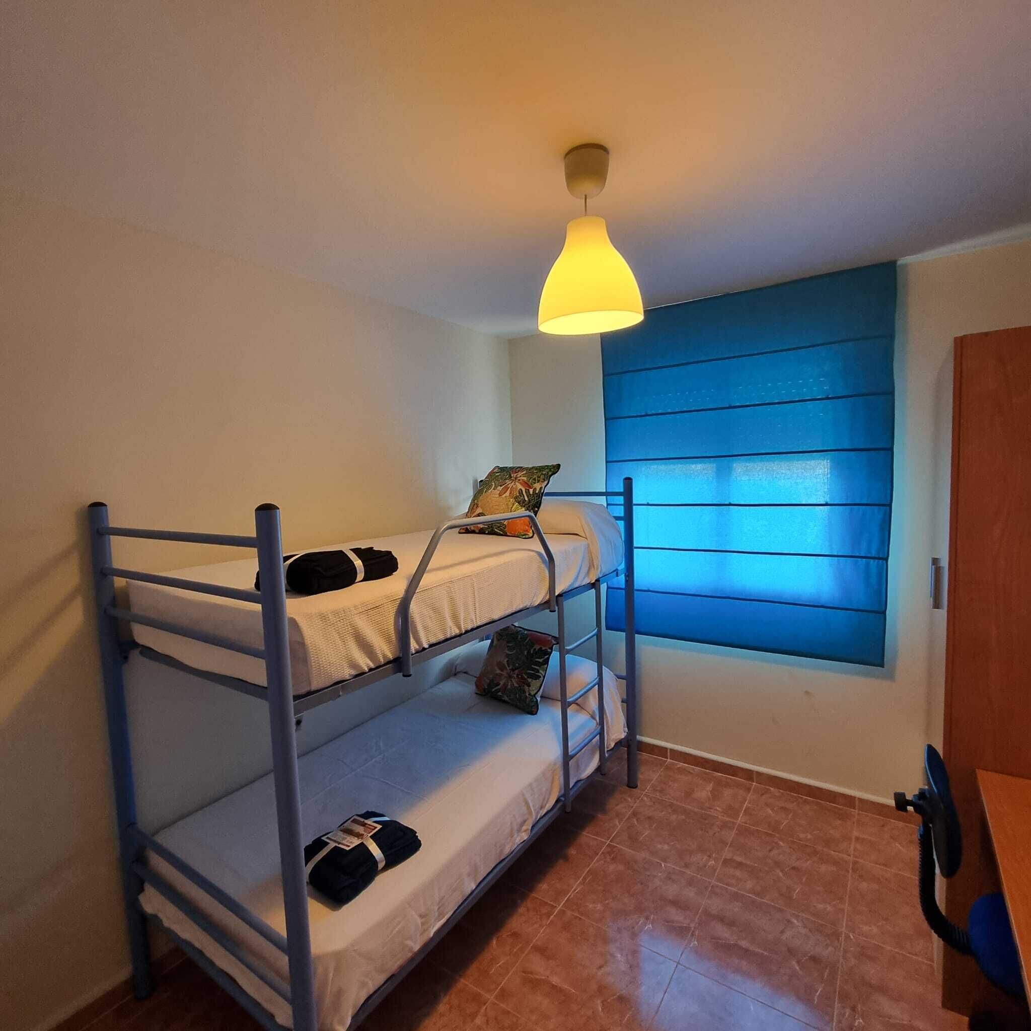 A dormitory room with bunk beds, a window with blue blinds, and a ceiling light.
