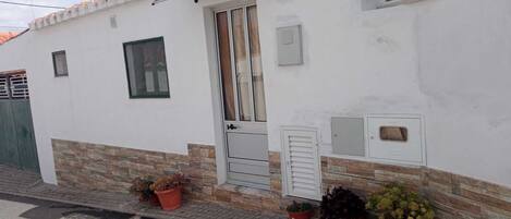 A white building with a tiled roof, a door, and windows. There are potted plants on the ground.