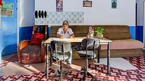A room with a brown sofa, a table with chairs, and a person writing at the table. - Hotel O Suci Homestay SyariahNearKilas 7 TV (Batam)