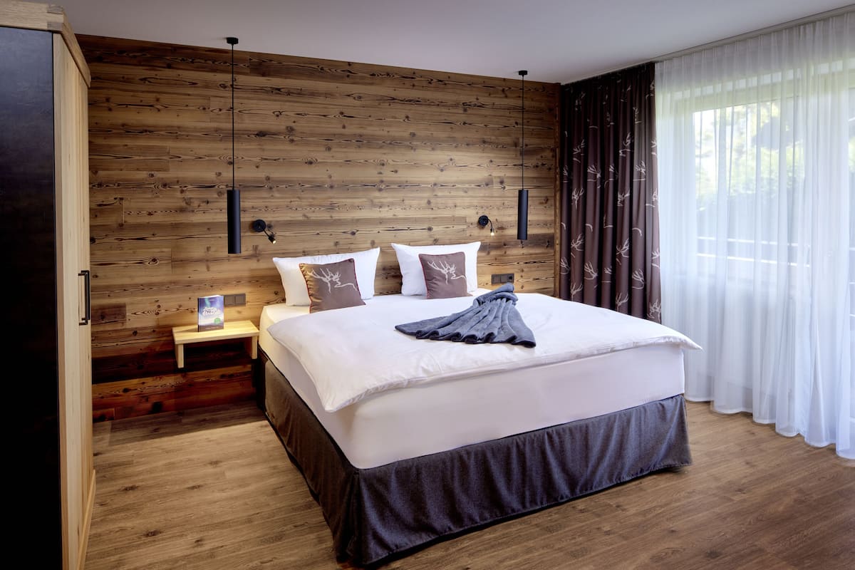 A bedroom with a wooden wall, a bed with white bedding, two bedside tables, and a window with sheer curtains.