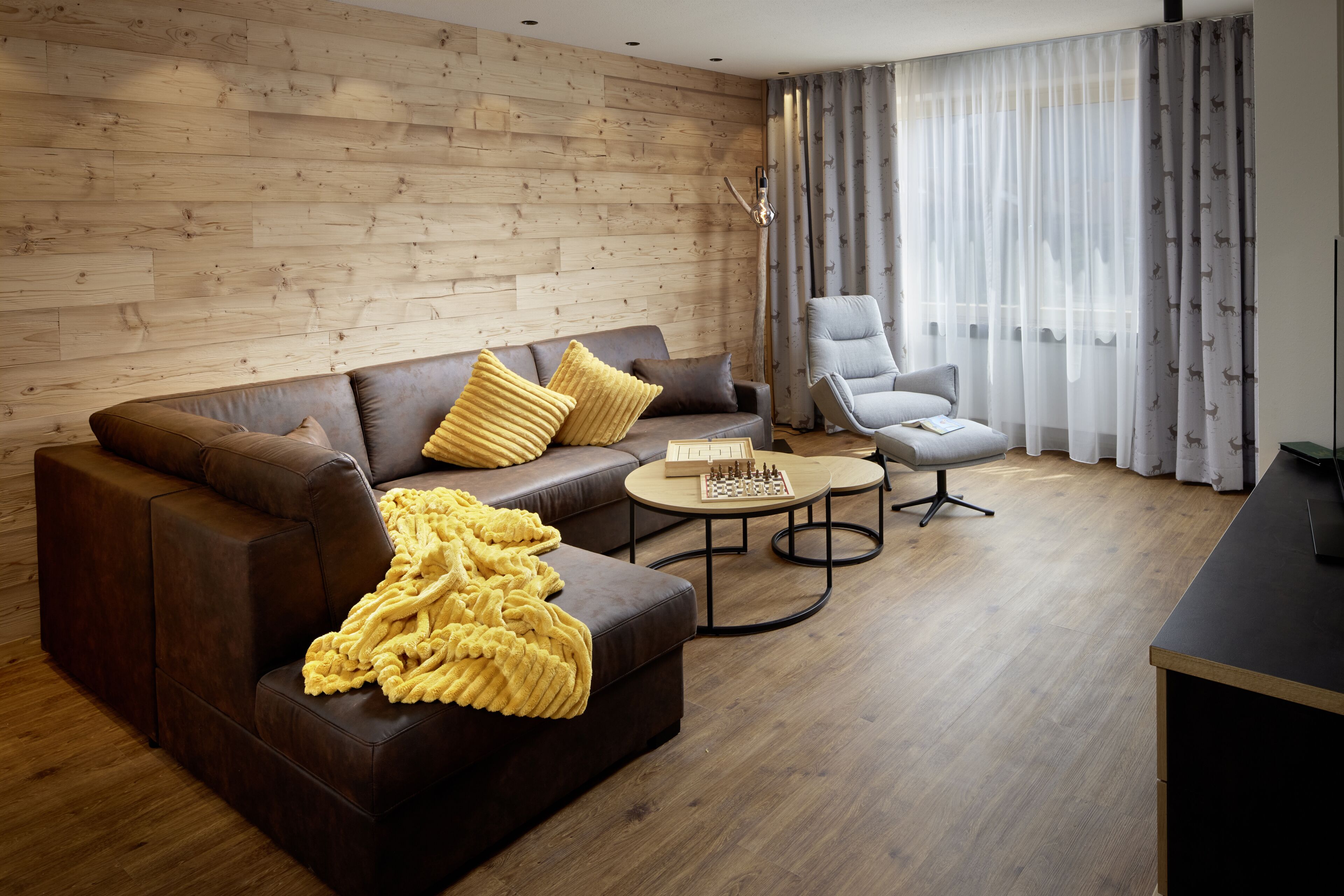 Un salon moderne avec un canapé en cuir marron, un plaid jaune, un fauteuil gris et une table basse ronde sur laquelle est posé un jeu d’échecs.