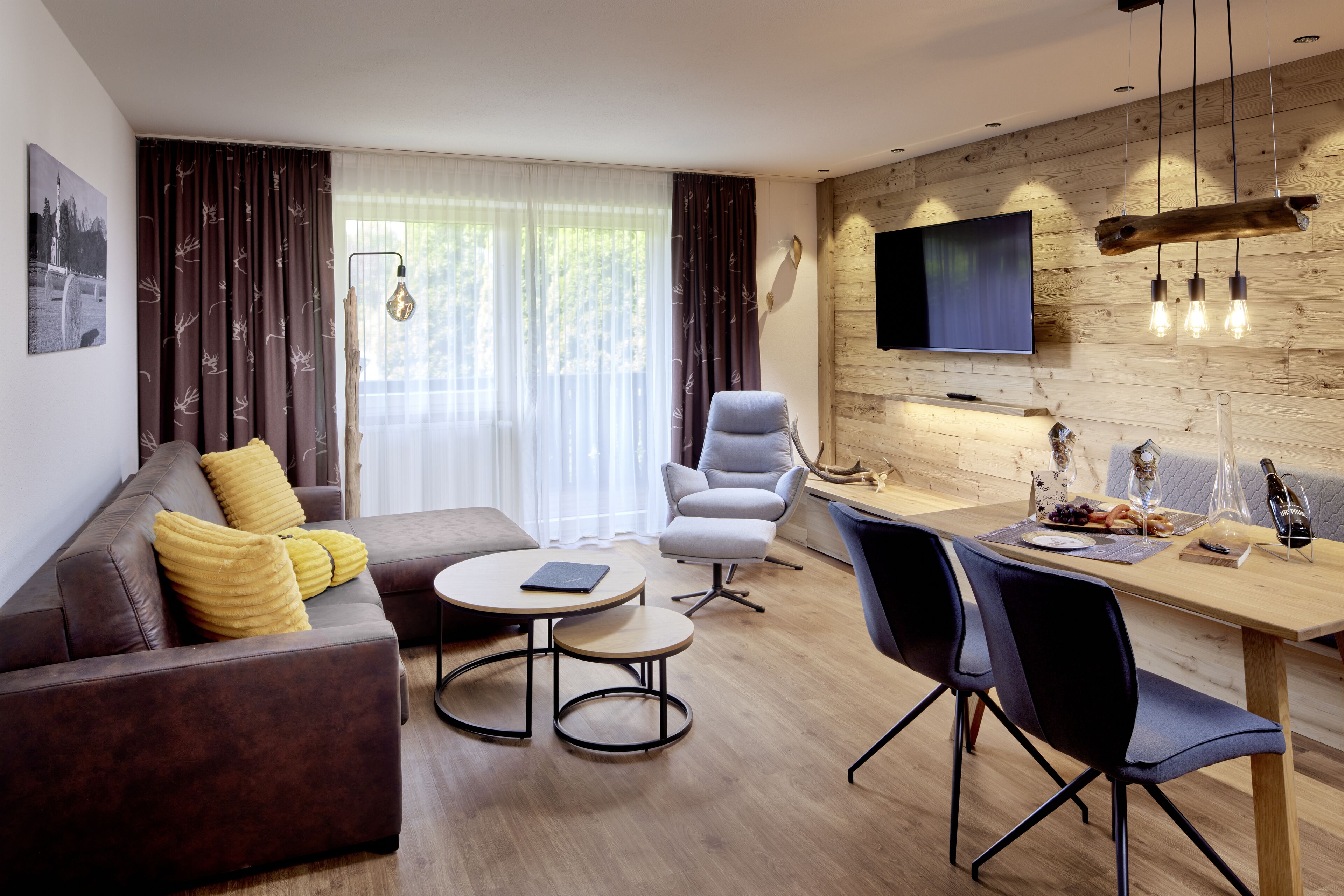 Ein modernes Wohnzimmer mit Sofa, Sessel, Esstisch und Fernseher an einer Holzwand.