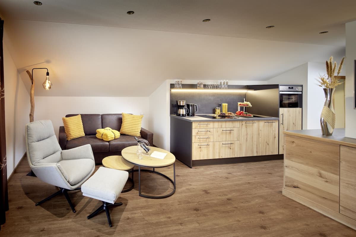A modern living room with a sofa, armchair, and coffee table, a kitchen with wooden cabinets and appliances, and a pendant light.