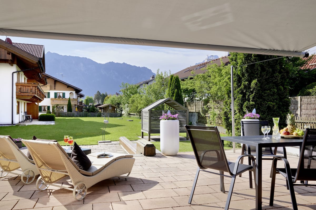 An outdoor patio with wicker chairs and a table, a white planter with flowers, and a view of a mountain.
