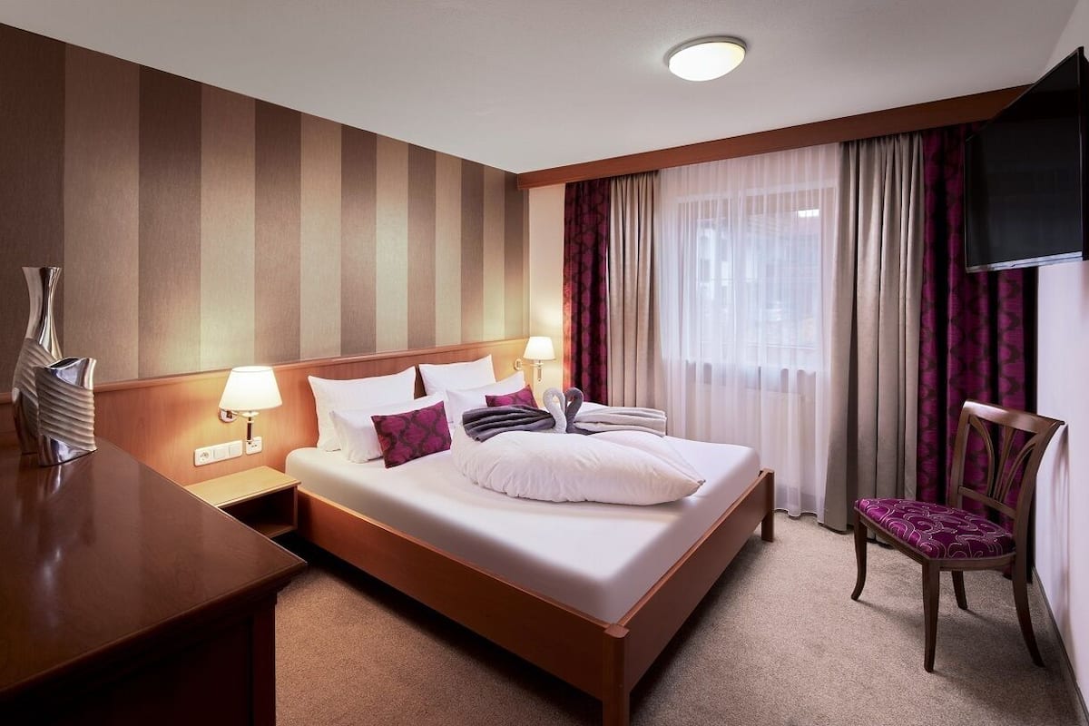 A hotel room with a bed, bedside tables, a chair, a TV, and curtains.