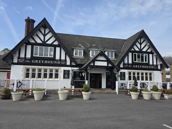 Un pub traditionnel appelé « The Greyhound », avec une façade noire et blanche, de nombreuses fenêtres et des tables à l’extérieur, agrémentées de plantes en pot.
