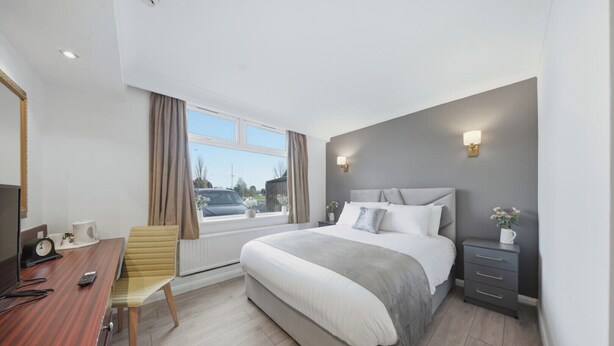 Una habitación de hotel moderna con una cama grande, un escritorio con silla, una mesita de noche con lámpara y una ventana con cortinas.