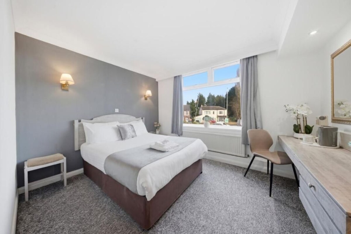 A hotel room with a bed, bedside table, chair, desk, and a window with a view of buildings.
