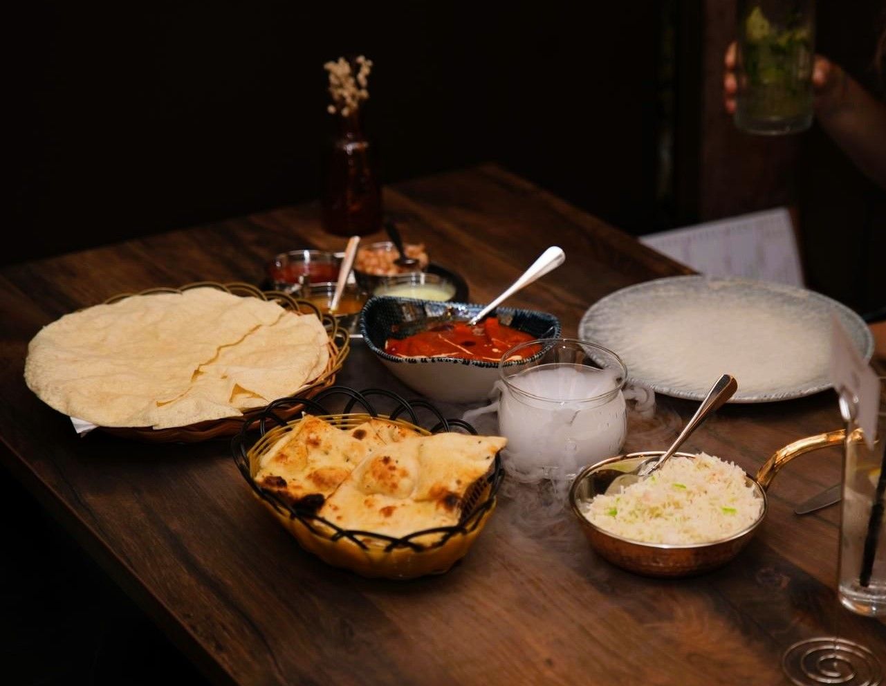 A wooden table with various food items including flatbread, naan, rice, and a bowl of sauce.