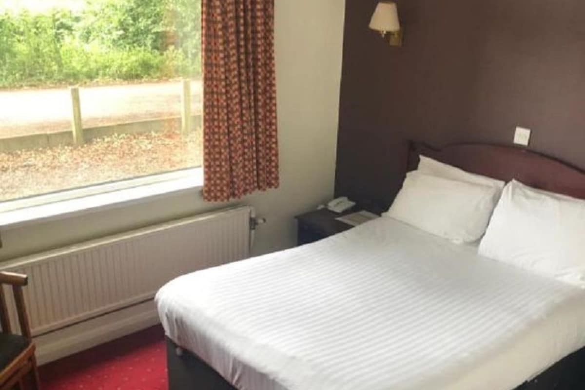A hotel room with a large bed, a nightstand, a chair, a window with curtains, and a radiator.