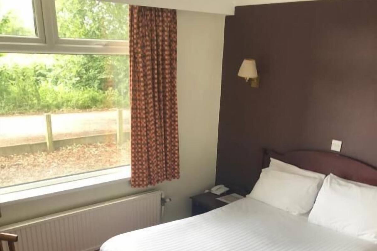 A hotel room with a large bed, a nightstand, a window with curtains, and a radiator.