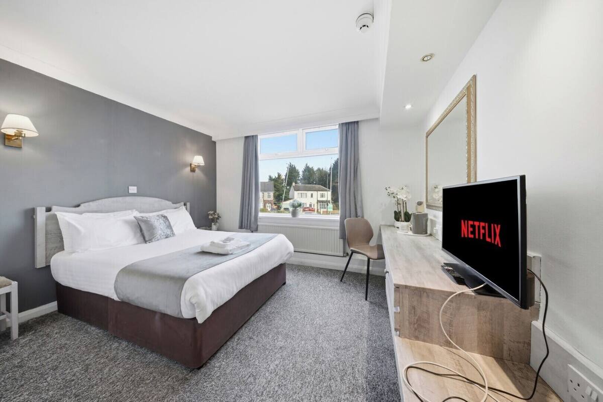 A modern hotel room with a large bed, a desk with a TV displaying the Netflix logo, and a view of buildings through the window.