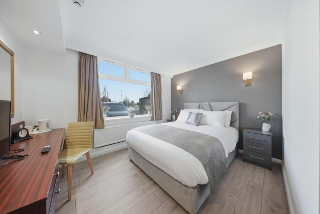 Una habitación de hotel moderna con una cama grande, un escritorio con una silla, una mesita de noche y una ventana con cortinas.