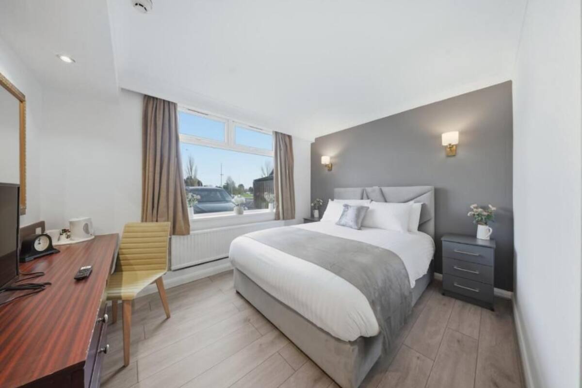 A modern hotel room with a large bed, a desk with a chair, a bedside table, and a window with curtains.