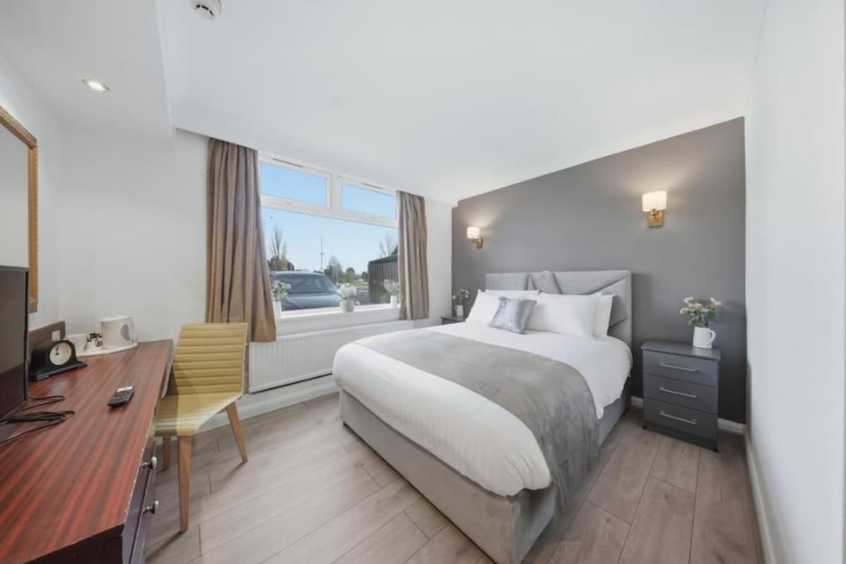 A modern hotel room with a large bed, a desk with a chair, a bedside table, and a window with curtains.