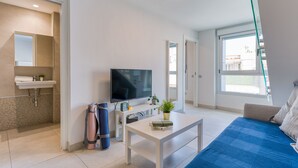 Un salon moderne avec un téléviseur à écran plat, une table basse blanche, un canapé bleu et une petite table blanche ornée d’une plante.