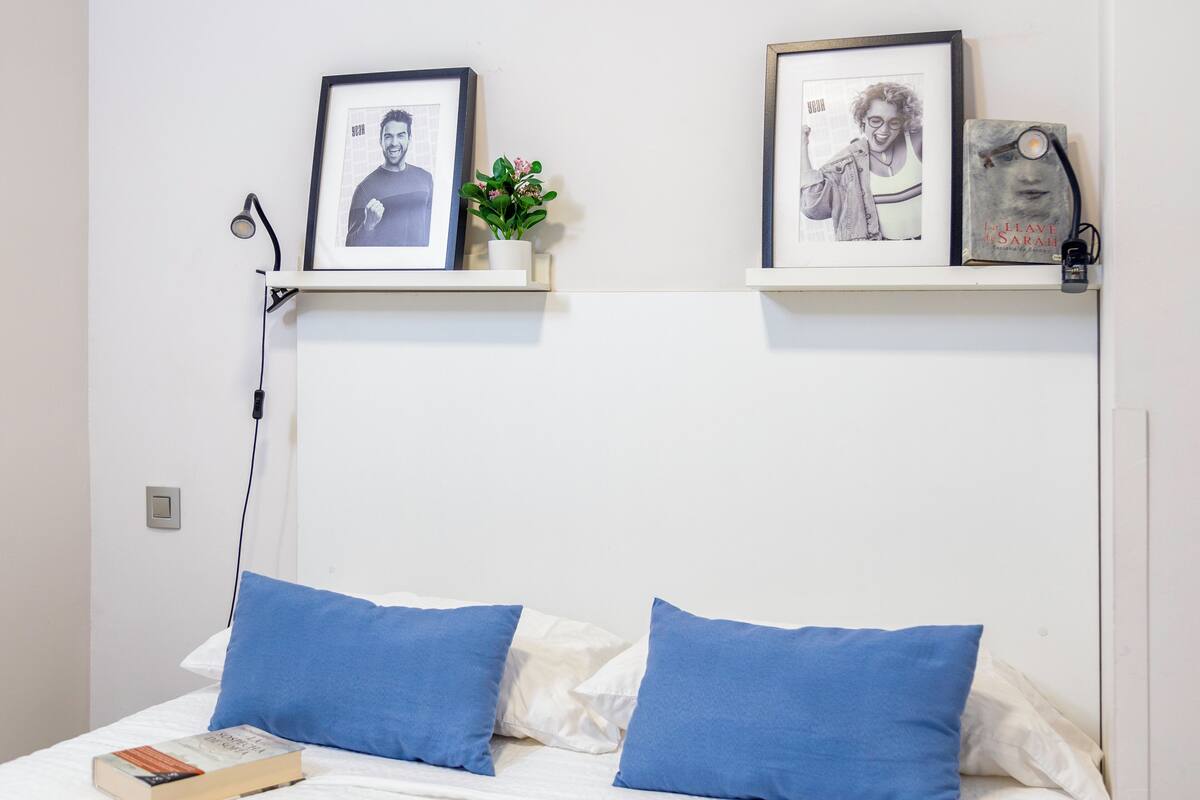 Cama bem arrumada com travesseiros azuis e brancos, um livro e duas fotografias emolduradas numa parede branca.
