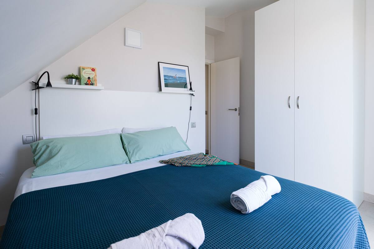 Quarto com cama, almofadas, mesa de cabeceira com abajur, quadro emoldurado e guarda-roupa.