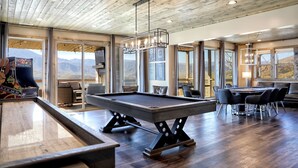 Una mesa de billar en una habitación con ventanas amplias, techo de madera y una zona de comedor con sillas y una mesa.