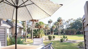 Un patio al aire libre con una mesa y sillas, un paraguas estampado y vista a un césped cuidado con palmeras.