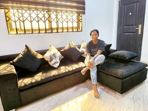 A person sitting on a dark-colored sofa with multiple cushions, a window with a metal grille, and a dark door. - Lovely & Cute Condo - Close to Highway - Parking (Amrahia)