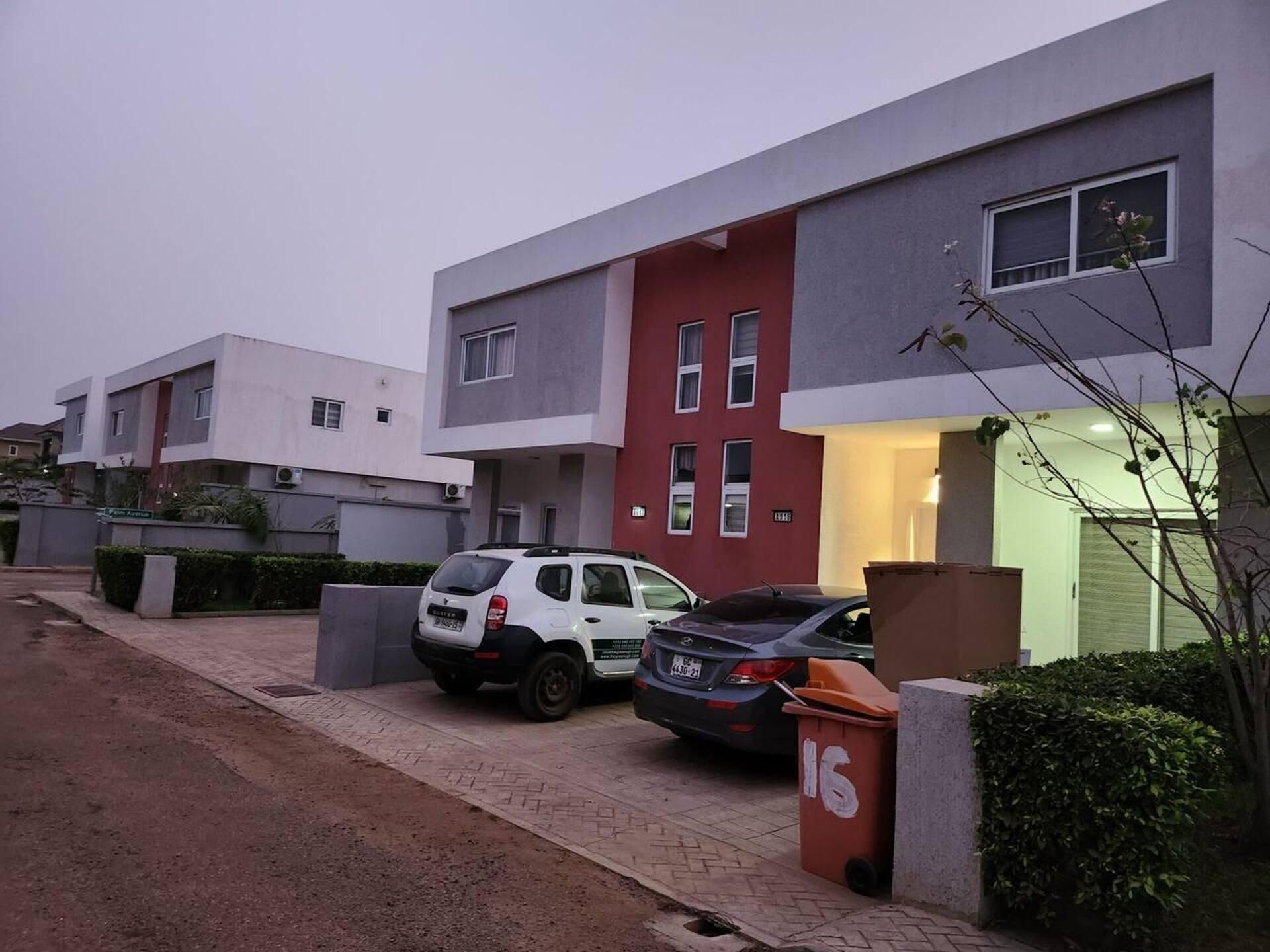 Un immeuble résidentiel moderne à la façade rouge et grise, avec des voitures garées et une poubelle.