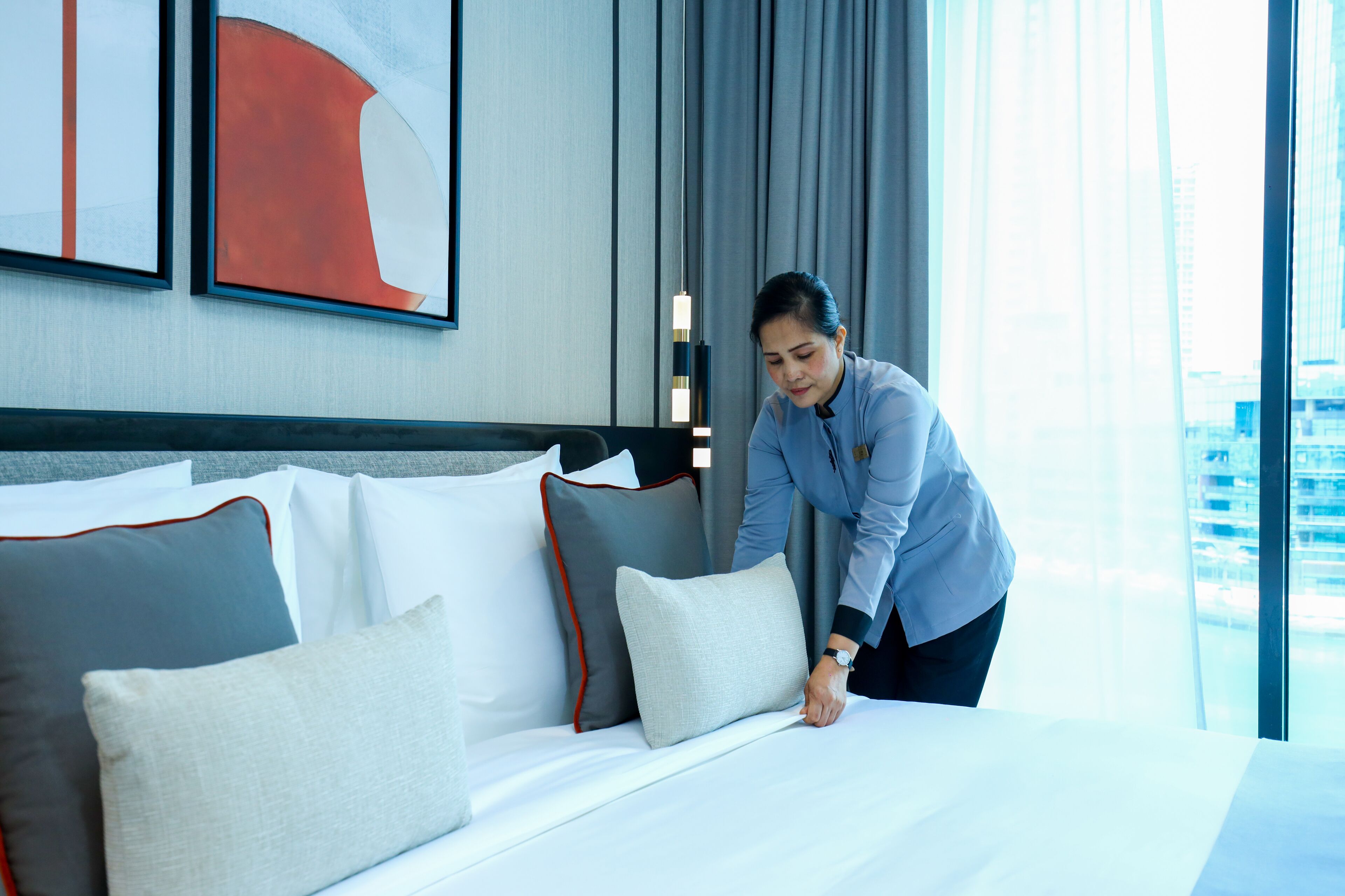 A hotel room with a neatly made bed, a large window with a view of buildings, and a modern abstract painting on the wall.