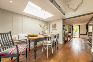 Un coin repas avec une table en bois, des chaises et un banc. Une lucarne se trouve au-dessus de la table, et un crâne de cerf est accroché au mur.