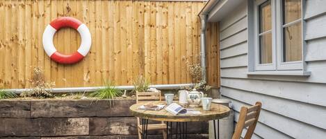 Una terraza de madera con una mesa redonda y sillas, un anillo salvavidas en la valla, y una ventana en la casa.