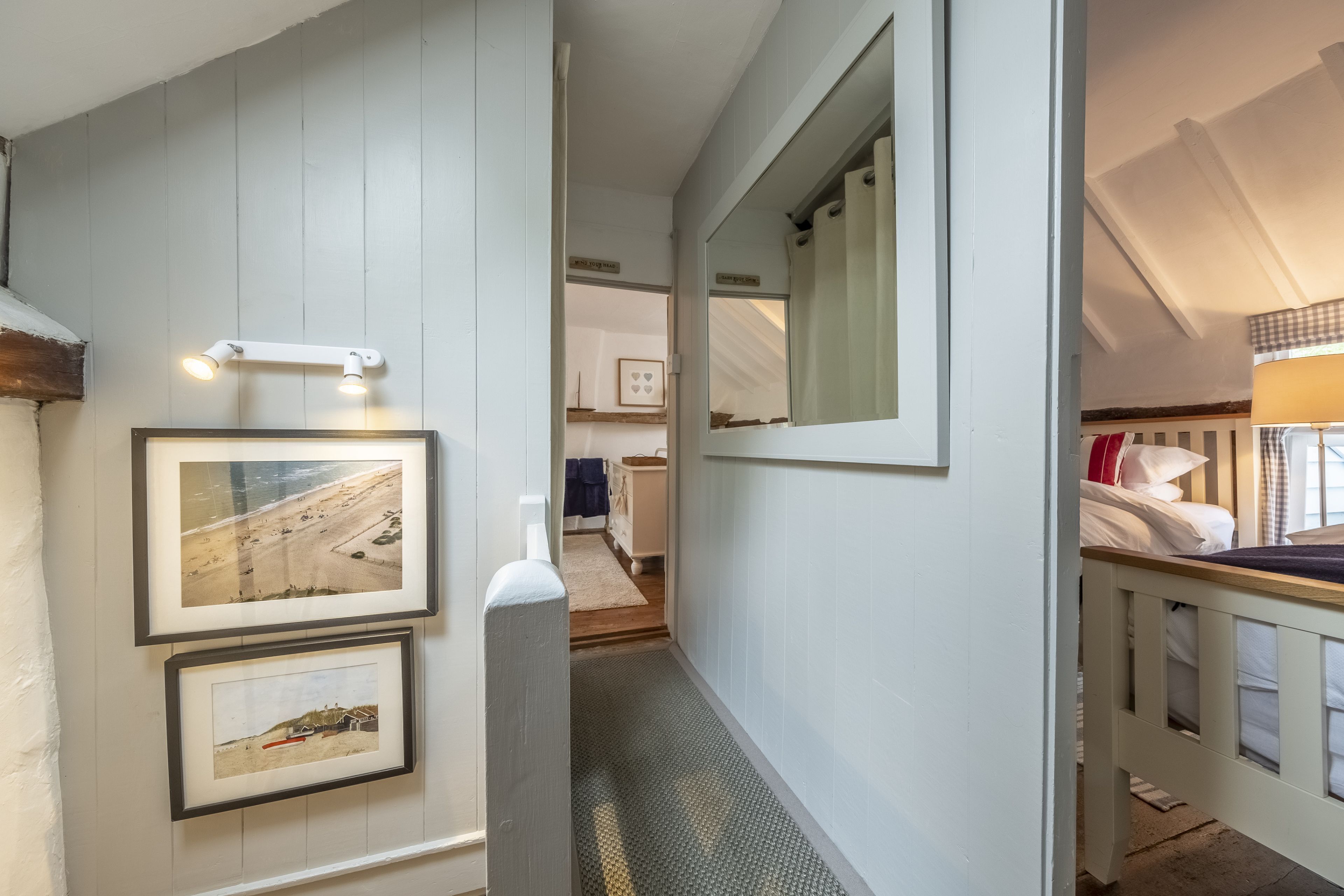 A hallway with framed pictures on the wall, a mirror, and a bed with a headboard in the adjacent room.