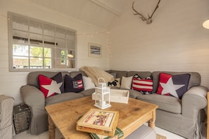 Un salon chaleureux avec une table basse en bois, un jeu de société Monopoly et une lanterne blanche. Un canapé orné de coussins aux couleurs du drapeau américain est adossé au mur, et une fenêtre illuminée de guirlandes lumineuses est visible.