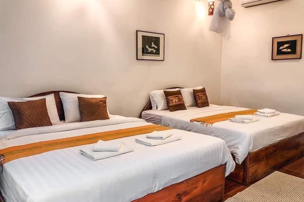 A hotel room with two single beds, each with a wooden headboard, white bedding, and brown decorative pillows. There are framed pictures on the walls and a small table with folded towels.