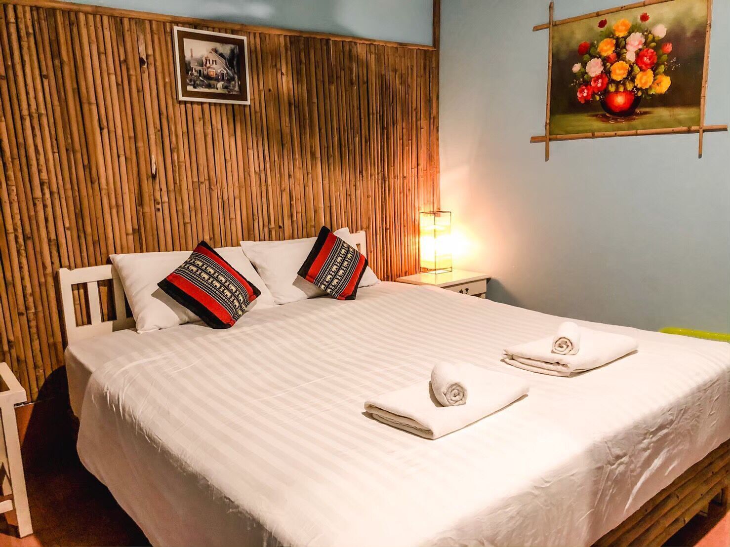 A neatly made bed with white linens and two pillows, a wooden headboard, a framed picture on the wall, and a painting of flowers.