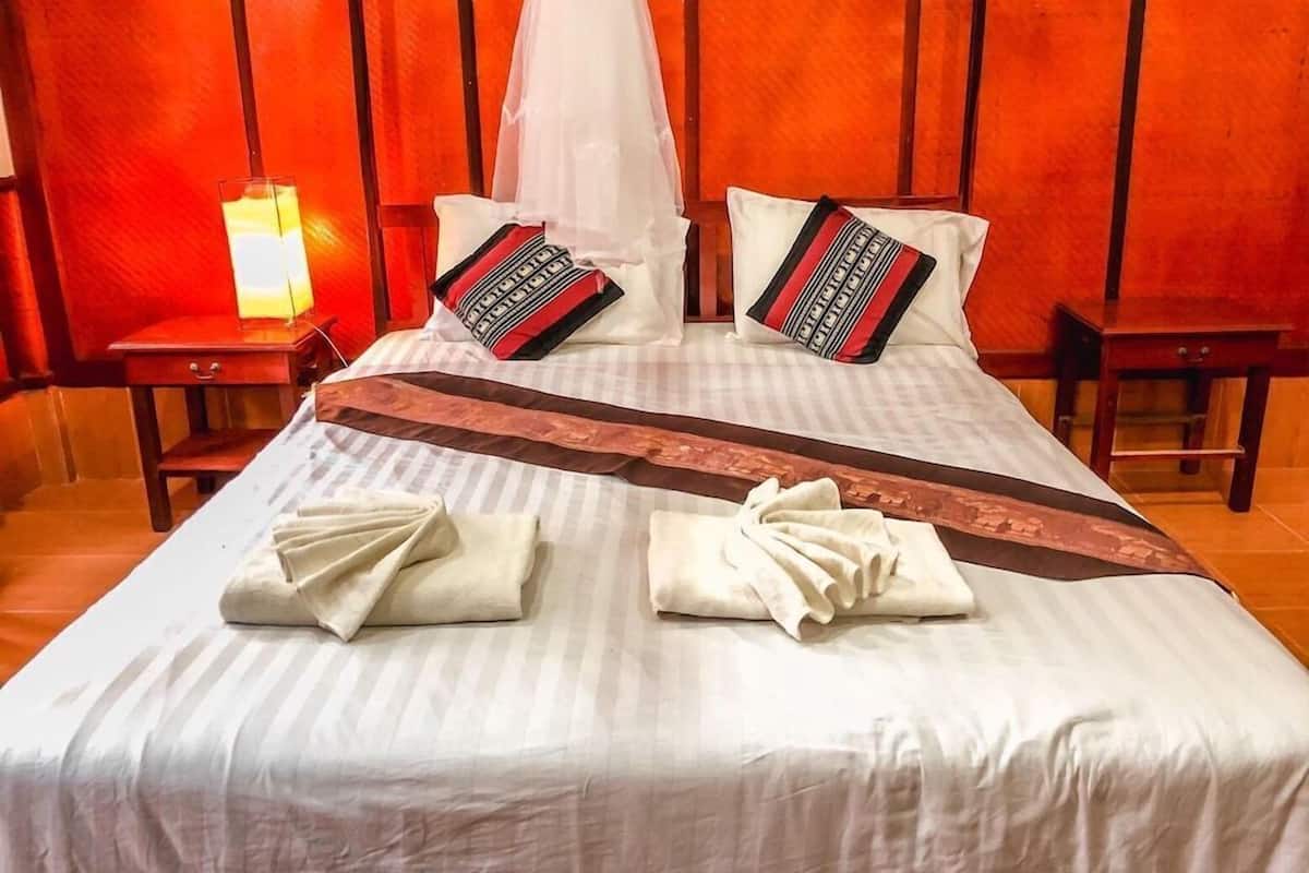 A neatly made bed with white linens, two pillows with red and black patterns, a brown decorative strip, and two folded towels.