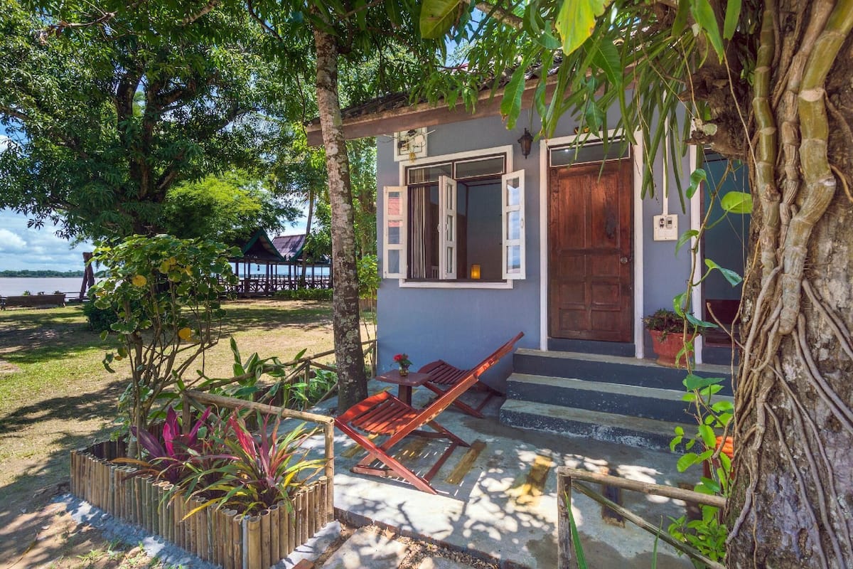 A small house with a wooden door and windows, a concrete porch, and a garden with plants and a wooden bench.