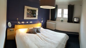 A neatly arranged hotel room with a large bed, bedside tables, a mirror, a picture on the wall, a window with curtains, and a radiator. - Ritterhof guest rooms at the Fitz-Ritter winery - VDP/Organic (Bad Dürkheim)