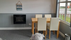 A modern living room with a grey sofa, a dining table with chairs, a fireplace, and a view through large windows. - Perfect Chalet to Relax in k4 (Mablethorpe)