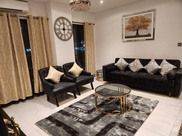 A modern living room with a black leather sofa, two armchairs, a glass coffee table, and a patterned rug. A large wall clock and a framed picture of a tree are also visible. - Luxurious & Comfy Gem-5 Star Location-pools, Gym! (Accra)