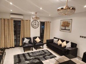 A modern living room with a black leather sofa, two armchairs, a large clock, and a chandelier. - Luxurious & Comfy Gem-5 Star Location-pools, Gym! (Accra)