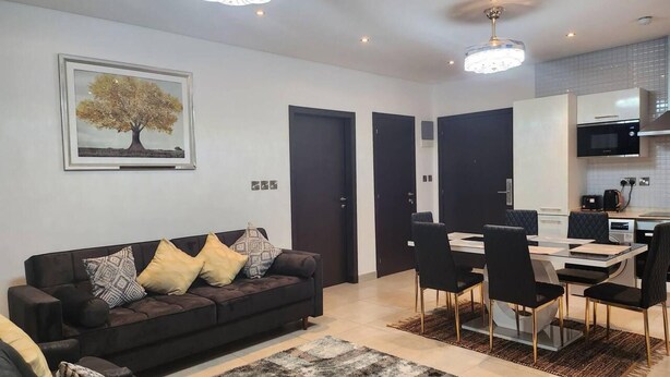 A modern living room with a dark-colored sofa, a dining table with chairs, and a kitchen area with cabinets and appliances. - Luxurious & Comfy Gem-5 Star Location-pools, Gym! (Accra)