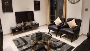 A modern living room with a flat-screen TV, a glass coffee table, two leather armchairs, and a large wall clock. - Luxurious & Comfy Gem-5 Star Location-pools, Gym! (Accra)