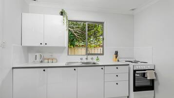 A modern kitchen with white cabinetry, a window with a view of trees, a kettle, a sink, and a stove.