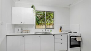 A modern kitchen with white cabinetry, a window with a view of trees, a kettle, a sink, and a stove.