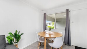 A dining area with a round table, white chairs, a potted plant, and a view through a window with curtains. - Beach Shack 5 (Crescent Head)