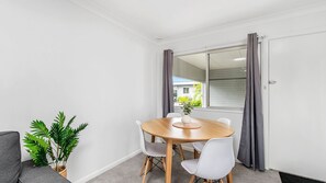 A dining area with a round table, white chairs, a potted plant, and a view through a window with curtains.